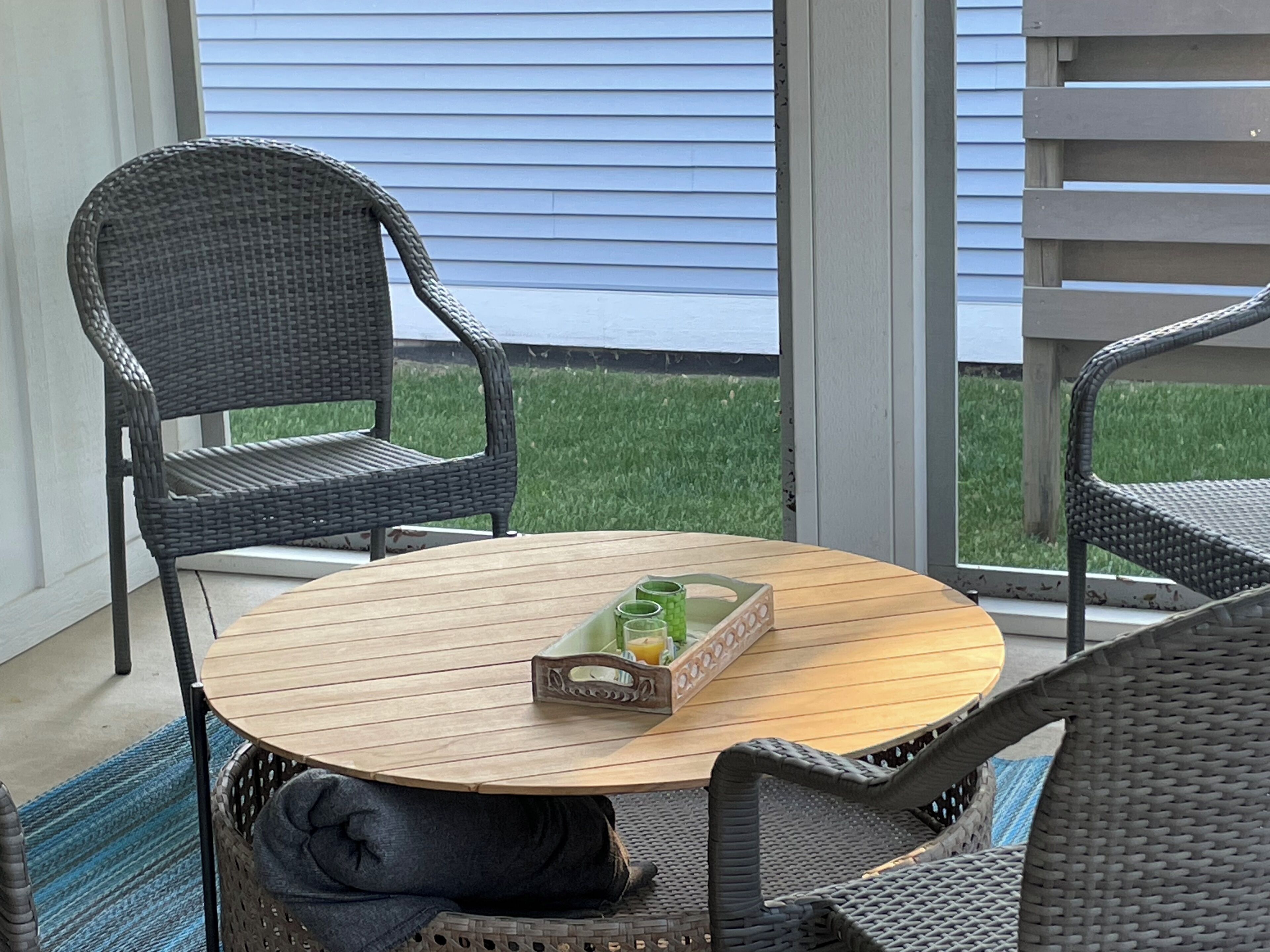 Une table ronde en bois, avec une chaise en osier de chaque côté, un panier en rotin contenant un plateau vert et une fenêtre avec des volets bleus.