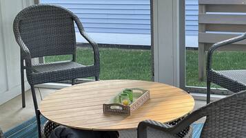 Une table ronde en bois, avec une chaise en osier de chaque côté, un panier en rotin contenant un plateau vert et une fenêtre avec des volets bleus.