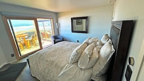 A bedroom with a large bed, a TV, a chair, and a sliding glass door leading to a balcony with a view of the beach.
