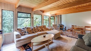 A spacious living room with a wooden ceiling, large windows, a sofa, a coffee table, and a dining area. - A 3 Story, 3 Bedroom Traditional Rustic Ski and Hiking Chalet (Deming)