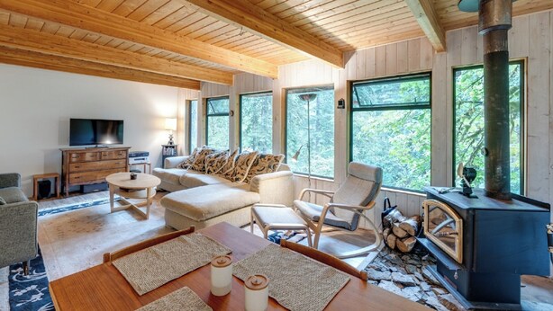 A cozy living room with a fireplace, wooden ceiling, and large windows offering a view of the outdoors. - A 3 Story, 3 Bedroom Traditional Rustic Ski and Hiking Chalet (Deming)