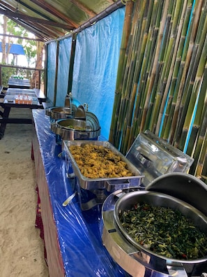 A buffet setup with multiple food service stations under a covered area with bamboo walls.