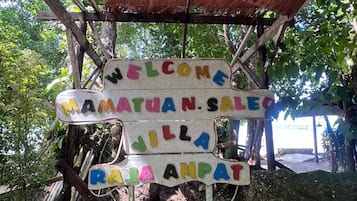 Une structure en bois surmontée d’une enseigne indiquant « BIENVENUE, VENTE MAMAYUAN, JILLA RAJA AMPAT », le tout entouré de verdure naturelle et d’éléments rustiques.