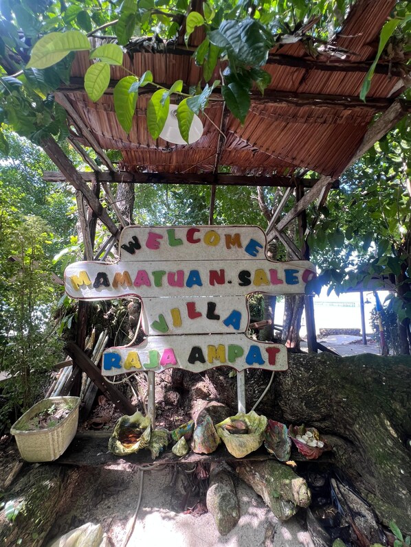 A wooden structure with a signboard displaying 'WELCOME MAMAYUAN SALE JILLA RAJA AMPAT' surrounded by natural foliage and rustic elements.