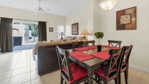 Un comedor con una mesa dispuesta para cuatro personas, rodeada de sillas con cojines rojos. Se ve un ventilador de techo sobre la mesa, y una lámpara de pared cuelga encima.