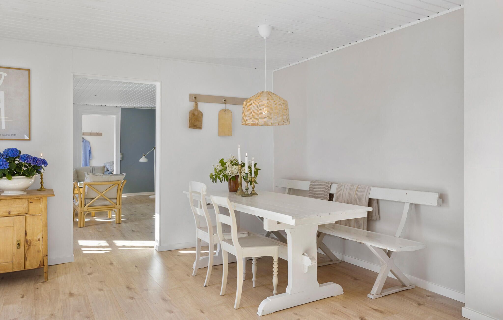 A dining area with a white table and bench, wicker pendant light, and a view into another room with a blue wall and a hanging garment.