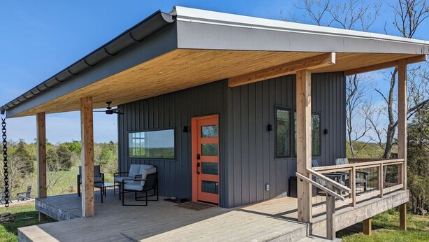 Une cabane moderne dotée d’une terrasse en bois, d’un espace salon extérieur abrité et d’une porte d’un orange vif.