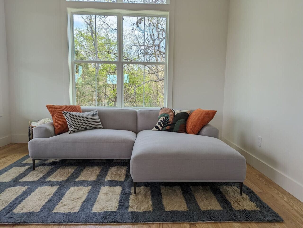 Una sala de estar moderna con un sofá modular gris, una alfombra estampada y un amplio ventanal con vistas a árboles.