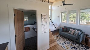 A modern interior with a staircase leading to a loft bedroom, a living area with a sofa and a ceiling fan, and a kitchen visible through an open door. - Your Own Two Cabin Retreat (Frankfort)