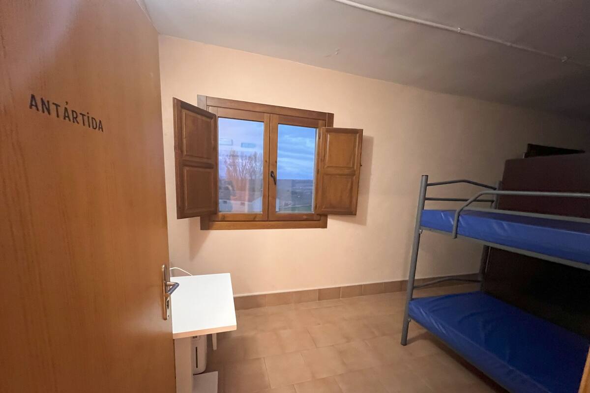 A room with a bunk bed, a small desk with a chair, a window with wooden shutters, and a door labeled "ANTARTIDA".