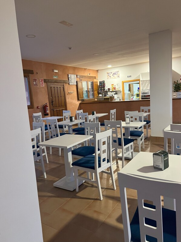 A restaurant with white tables and chairs, a bar area, and a fire extinguisher on the wall. - Ikigai Albergue  (Navarrete)