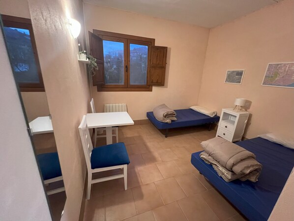 Une petite pièce bien éclairée, avec une table blanche, deux lits simples, une chaise blanche, un petit meuble et une fenêtre donnant sur des immeubles.