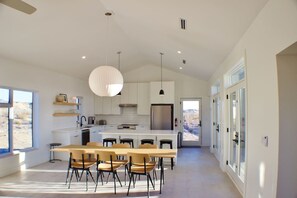 A modern kitchen with a central island, pendant lighting, and a view of the desert through the door.