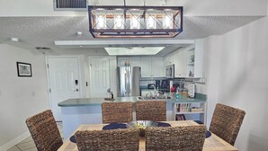 A dining area with wicker chairs, a wooden table, and a kitchen in the background. - Beach Condo at John's Pass (Madeira Beach)