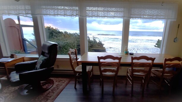 A dining area with a view of the sea, a table, chairs, and a window with lace curtains.