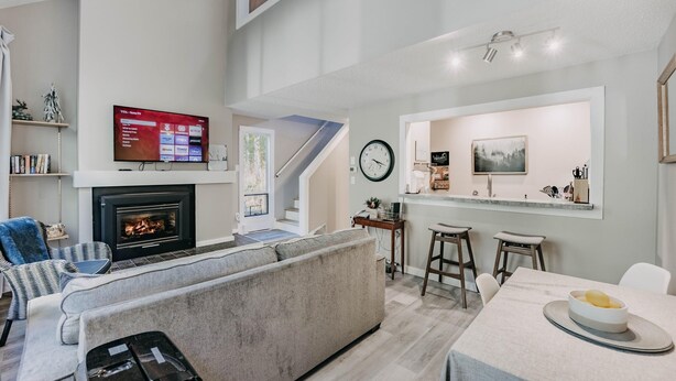 A modern living room with a fireplace, a TV mounted on the wall, a staircase leading to an upper level, and a dining area with stools. - Snowater Condo 45sw With a Media Room (Deming)