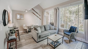 A modern living room with a grey sofa, a coffee table, a wicker chair, and a large window offering a view of trees. - Snowater Condo 45sw With a Media Room (Deming)