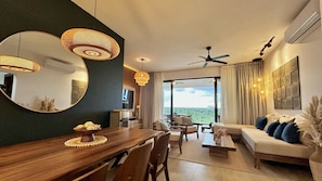 A modern living room with a wooden dining table, comfortable seating, a large mirror, and a view of the outdoors.