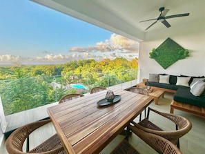 A balcony with a wooden table and wicker chairs, overlooking a lush landscape with trees and a pool.