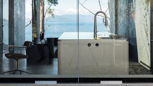 A modern interior with a large window overlooking a body of water, a central seating area, and a built-in kitchen with a sink. - Vipp Tunnel - Tasmania (Apollo Bay)
