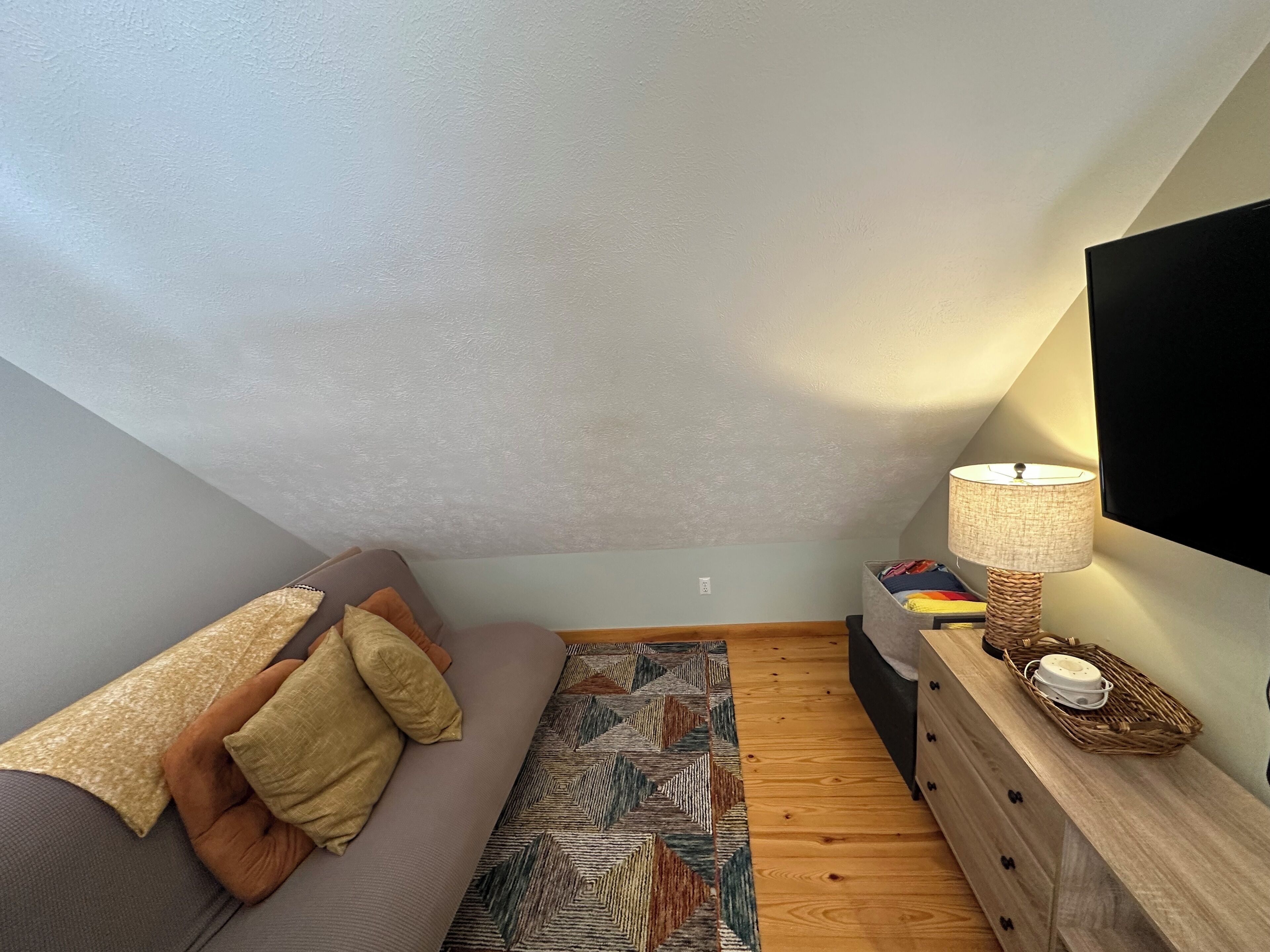 A cozy loft bedroom with a sloped ceiling, a sofa with patterned cushions, a wooden dresser, and a lamp.