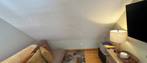 A cozy loft bedroom with a sloped ceiling, a sofa with patterned cushions, a wooden dresser, and a lamp.