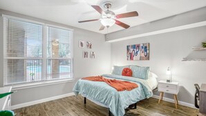 Un dormitorio con una cama, una mesita de noche, un ventilador de techo, una ventana con persianas y una obra de arte en la pared.