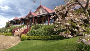 Uma casa grande e tradicional com telhado vermelho, várias varandas e um jardim bem cuidado com árvores floridas.