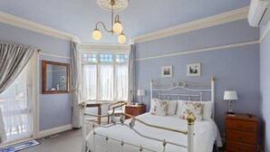 Quarto com cama branca, mesa de cabeceira de madeira, cômoda, escrivaninha e cadeira. Há uma janela com cortinas, um espelho e um candelabro.