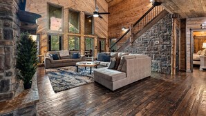 A spacious living room with a stone fireplace, large windows, and a wooden ceiling.