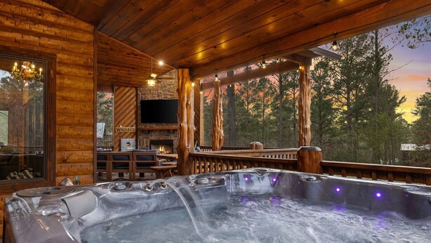 A wooden cabin interior with a hot tub, stone fireplace, and a view of the forest.