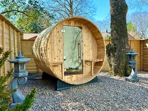 Una estructura en forma de barril de madera, con una puerta, rodeada de grava y árboles.
