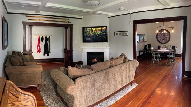 A living room with a fireplace, a television, and a dining area with a chandelier. - Heritage Apartment w City and Mountain Views (North Vancouver)