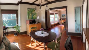 A dining area with a wooden table, chairs, and a vase with flowers. There is a window with blinds, a door, and a painting on the wall. - Heritage Apartment w City and Mountain Views (North Vancouver)