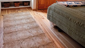 Una camera da letto con un armadio in legno, un letto verde, un tappeto intrecciato, una finestra con una pianta e uno scaffale con scatole portaoggetti.