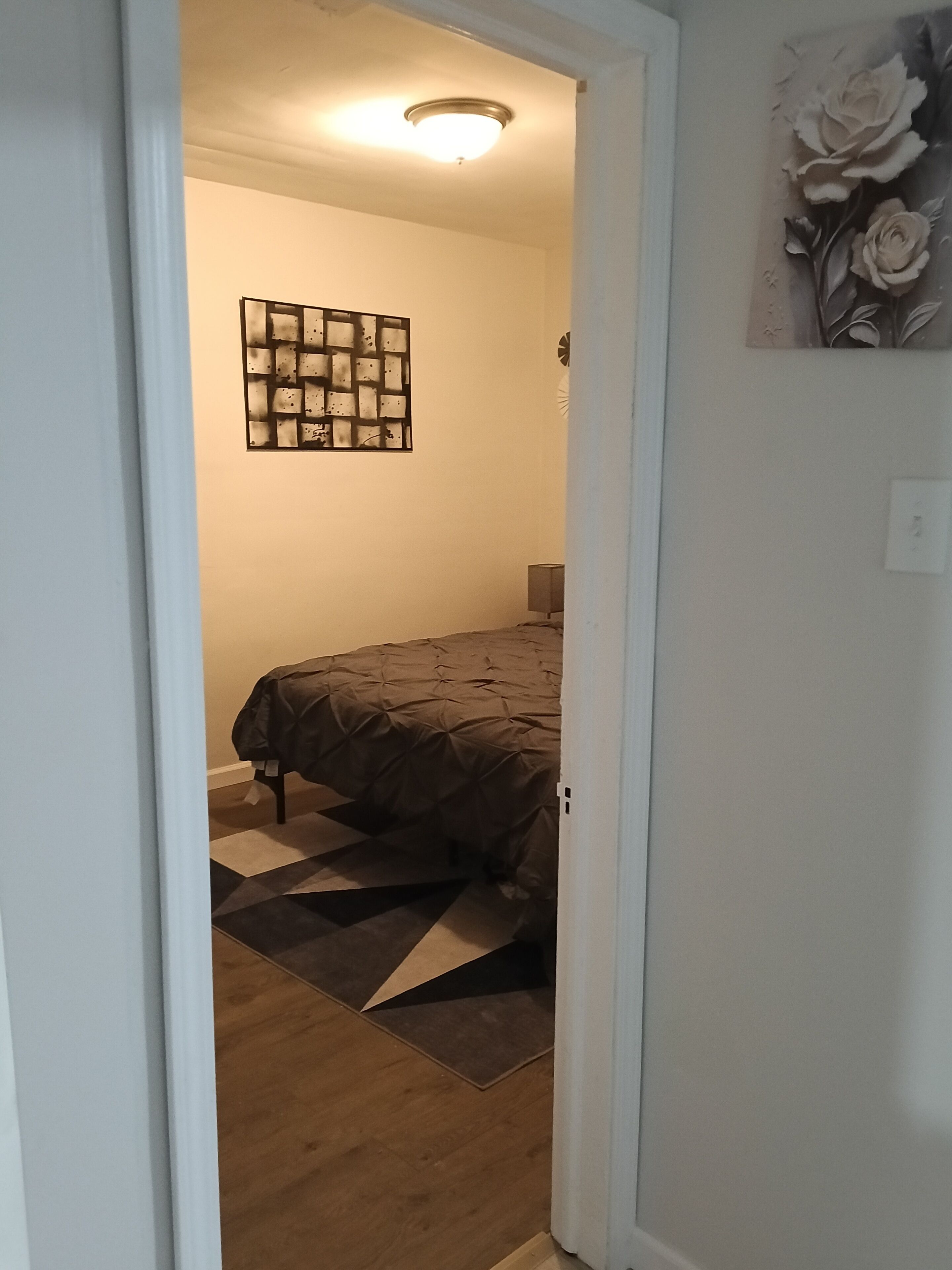 A bedroom with a bed, a geometric rug, a wall-mounted light fixture, and a floral artwork.