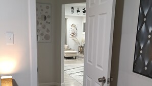 Un couloir avec une porte blanche, une table de chevet noire surmontée d’une lampe, un tapis moelleux et un mur orné de motifs circulaires.