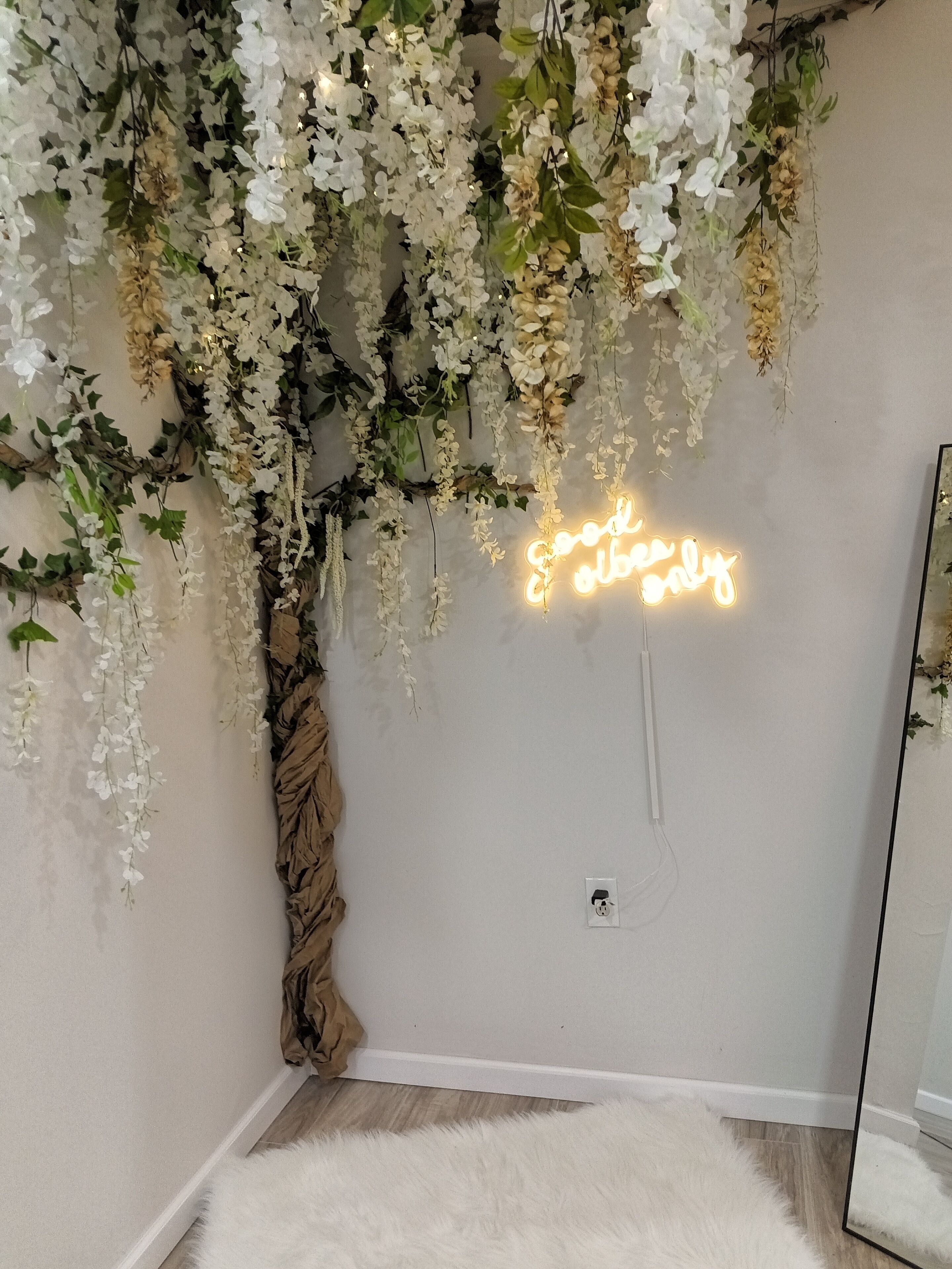 A room with a white shaggy carpet, a wall-mounted neon sign, and hanging floral decorations.