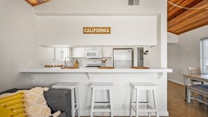 Cocina compacta con muebles blancos, un microondas, un refrigerador y un letrero que dice 'CALIFORNIA' sobre la encimera. Hay tres taburetes blancos y un sofá gris con una almohada amarilla.