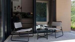 Un espace extérieur moderne aménagé avec deux chaises et une petite table, une porte vitrée donnant sur une cuisine, et une vue sur la verdure.