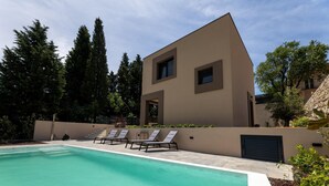 Une maison moderne à deux étages, dotée d’une piscine, entourée de grands arbres et d’un mur en pierre.
