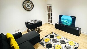 A living room with a black sofa, a yellow pillow, a black coffee table, a hexagonal patterned rug, a wall clock, and a television displaying a travel channel. - Ground Floor Entire Holiday Apartment - Self Check in/out - Perfect for 6 Guests (Crossgates)