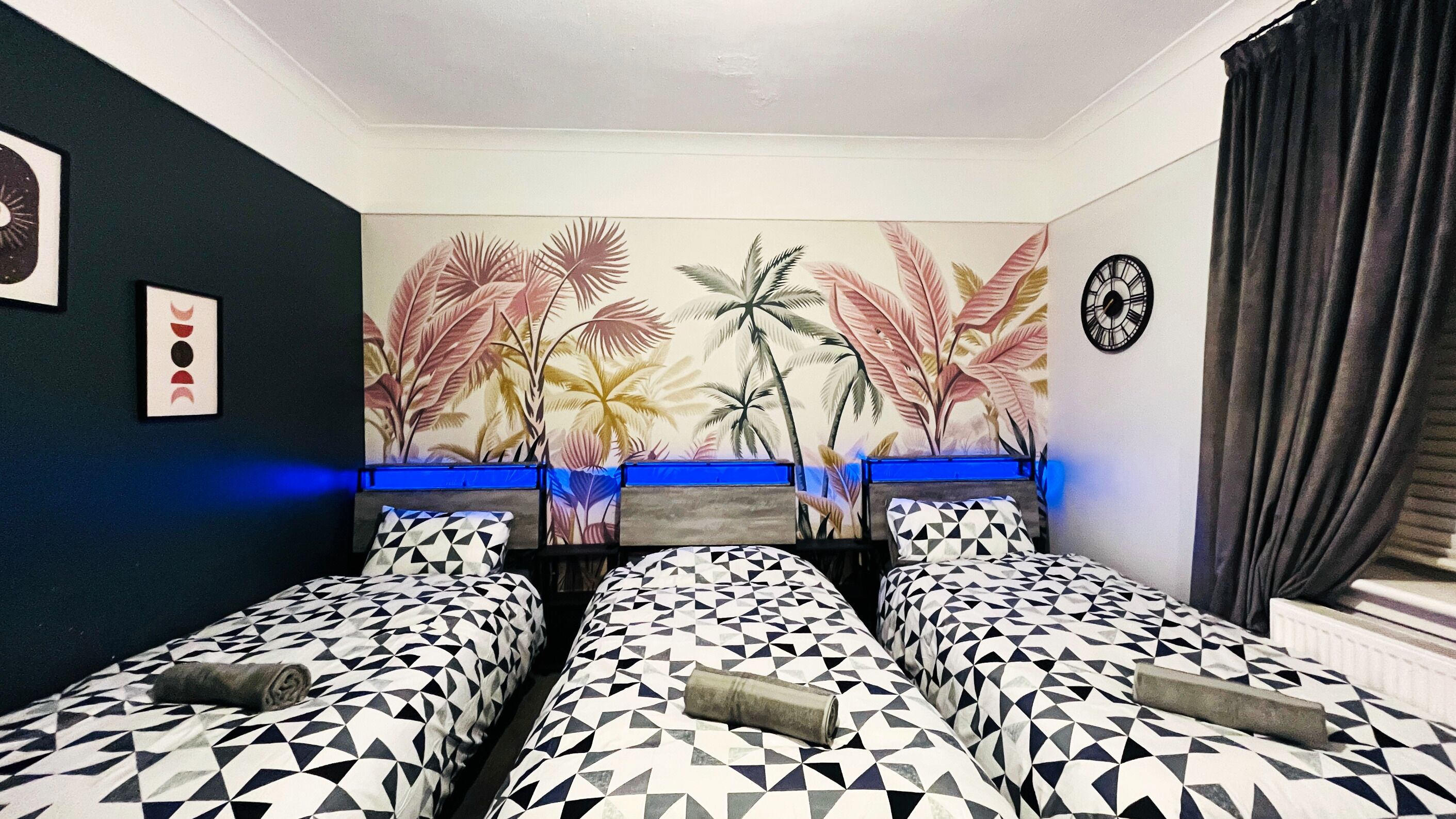 A room with two beds, a tropical-themed wall mural, a clock, and a window with curtains.