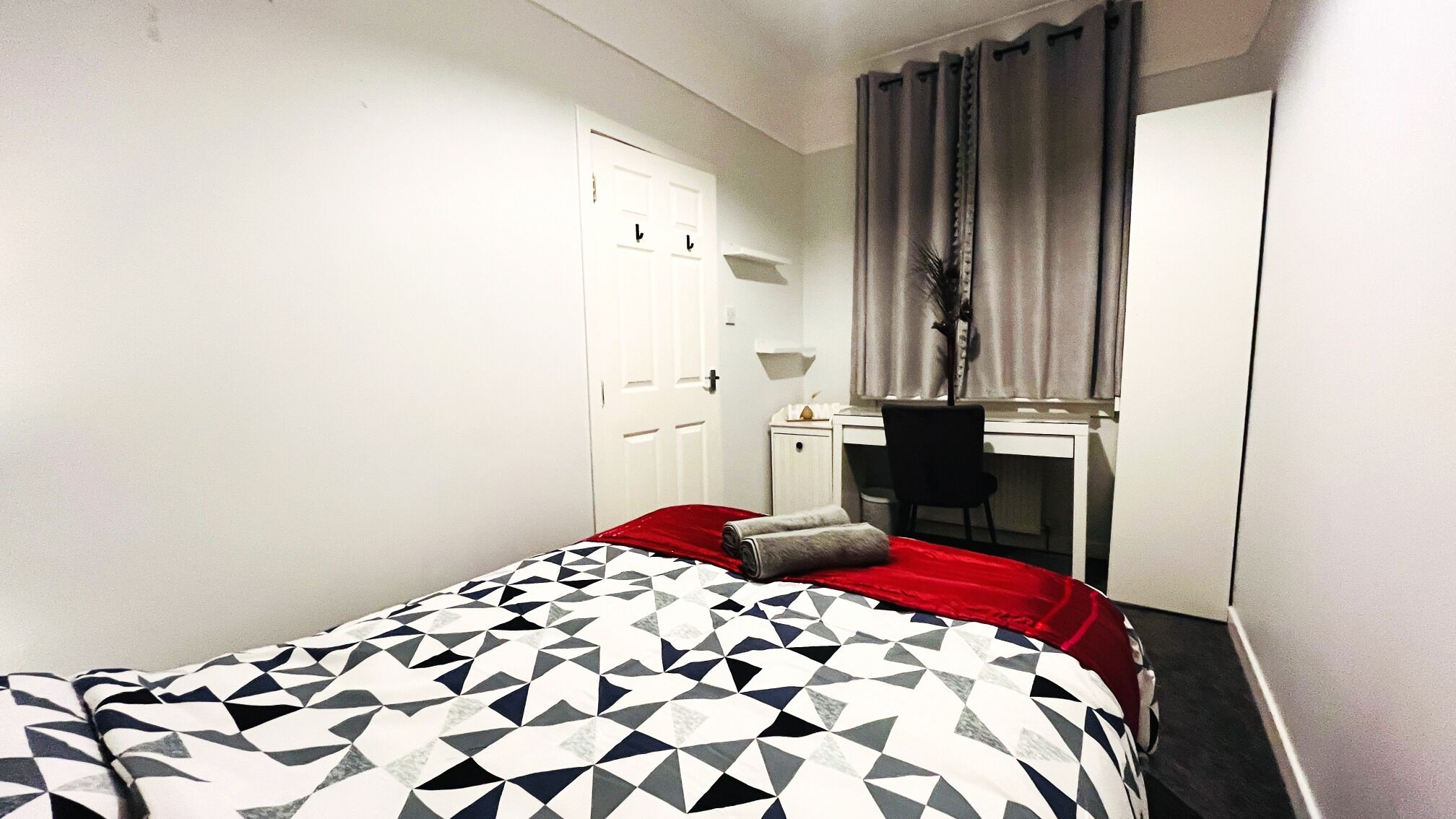 A bedroom with a bed, a desk, a chair, a wardrobe, and a window with curtains.