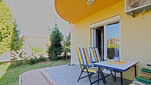 Un patio exterior con mesa y sillas, una mesita con una taza, y una ventana con vista a una casa y árboles.