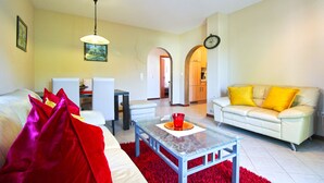 Una sala de estar con un sofá, una mesa de centro de vidrio, un sillón blanco, una alfombra roja, una mesa de comedor con sillas y una cocina visible a través de un arco.