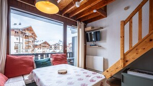 A cozy room with a wooden ceiling, a large window offering a view of buildings, a table with a patterned tablecloth, and a TV mounted on the wall. - Cosy mezzanine studio for 4 ppl (Morillon)