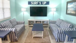 A living room with striped sofas, a coffee table, a TV, and a sign that reads "SURF'S UP".