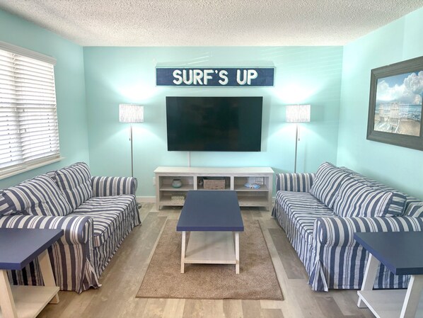 Una sala de estar con sofás a rayas, una mesita de centro, un televisor y un letrero que dice "SURF'S UP".