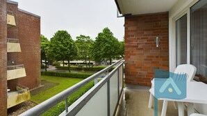 A balcony with a view of a green lawn and trees, a white table with a chair, and a brick building with a window. - In 2nd row to the beach - Gmelinstraße 8 (Wyk auf Föhr)